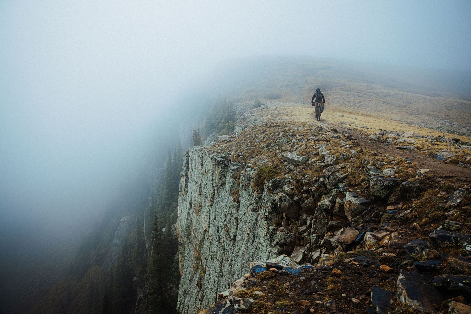 A lone cyclist stands at the edge of a misty cliff face in moody weather conditions.