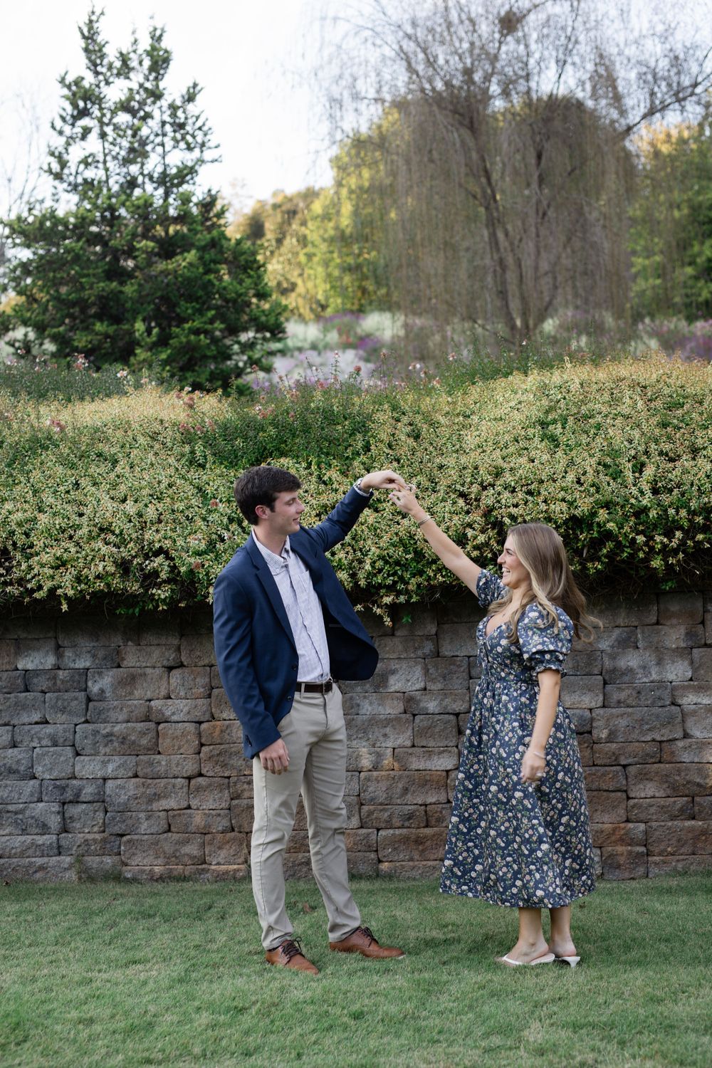 A couple dances playfully in a garden setting with stone wall backdrop.