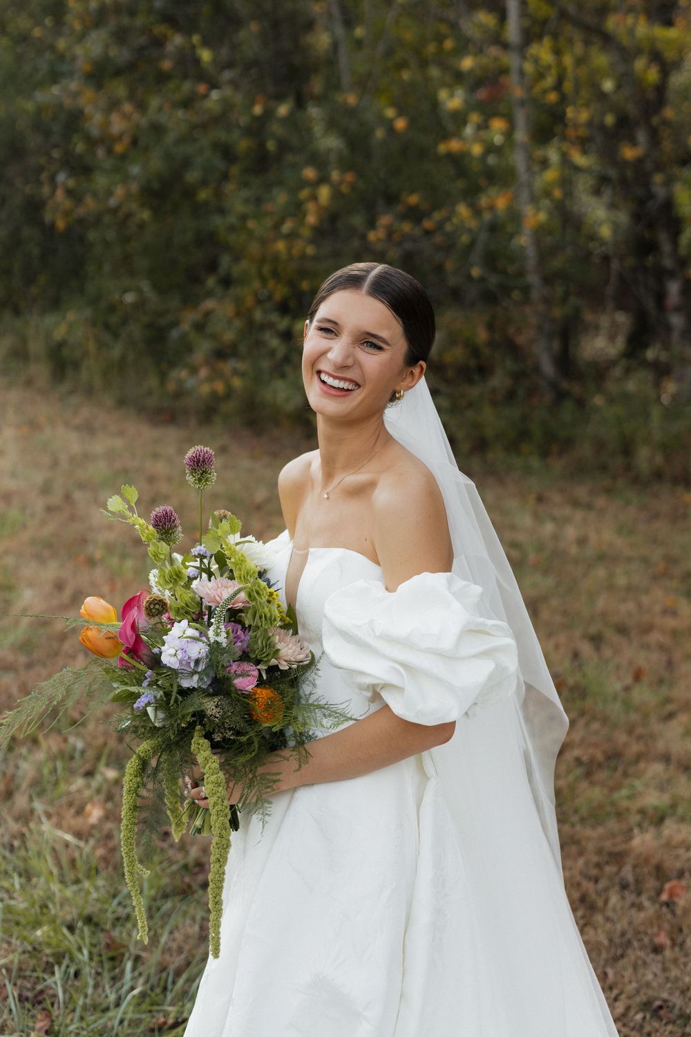 Bride in off-shoulder wedding dress holding colorful wildflower bouquet against autumn backdrop.