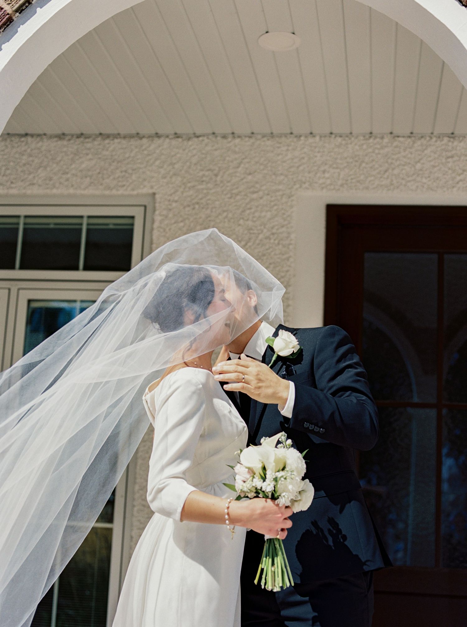 Bride and groom sharing a kiss under flowing veil outside chapel doors.
