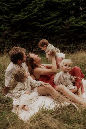 Joyful family sitting in tall grass, mother lifting baby while children gather close outdoors.