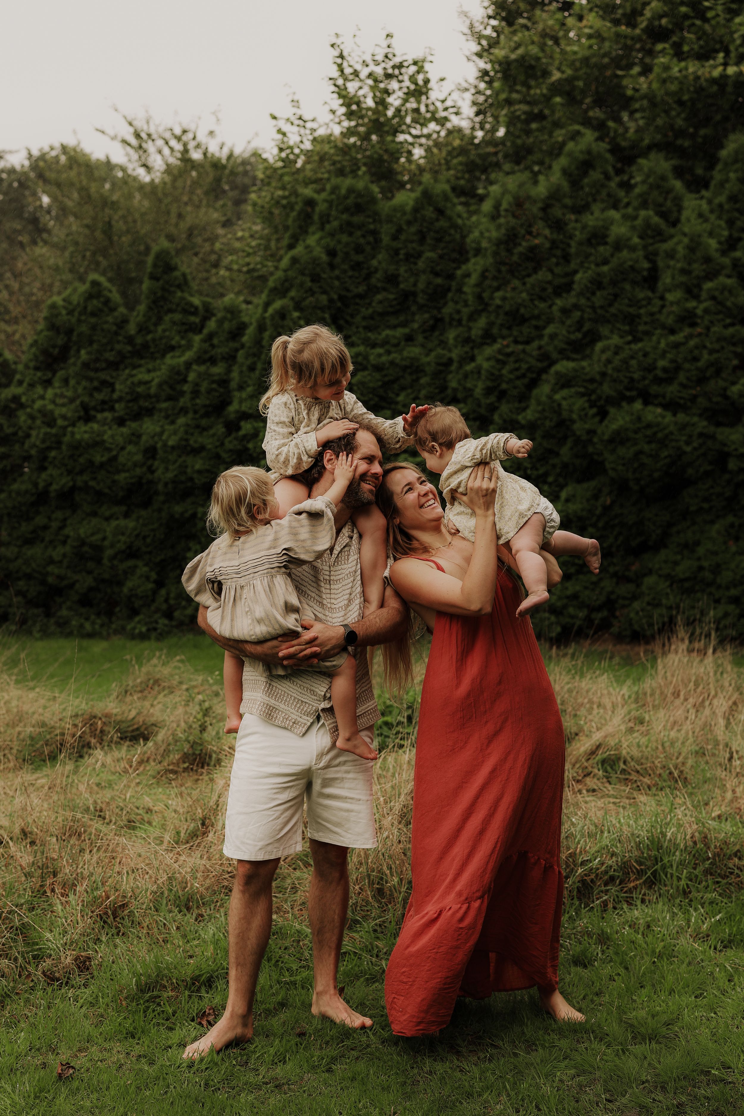 Happy family of five playing outdoors, parents lifting children in a lush green garden setting.