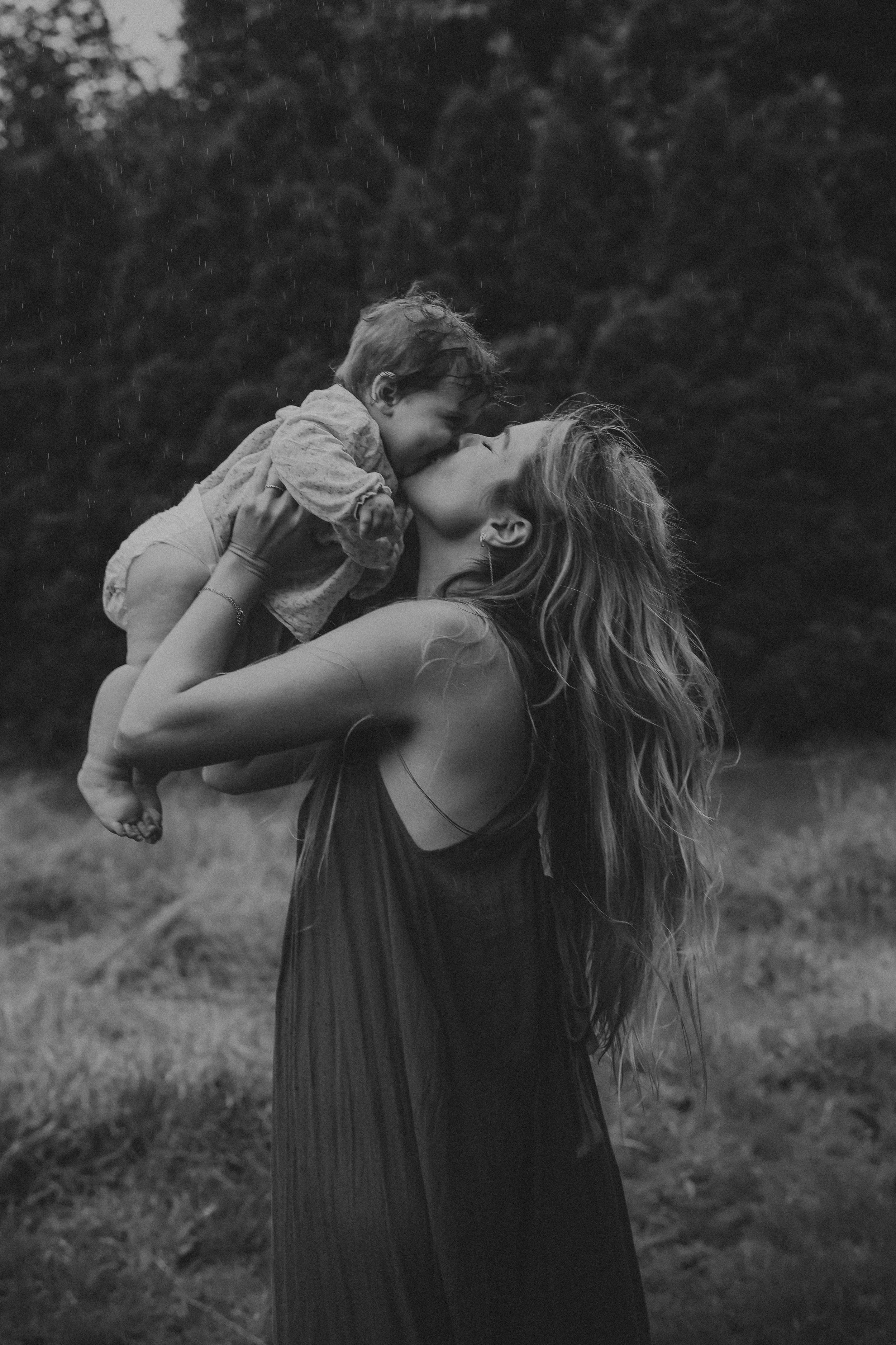 Black and white photo of a mother lifting and kissing her baby in an outdoor garden setting.
