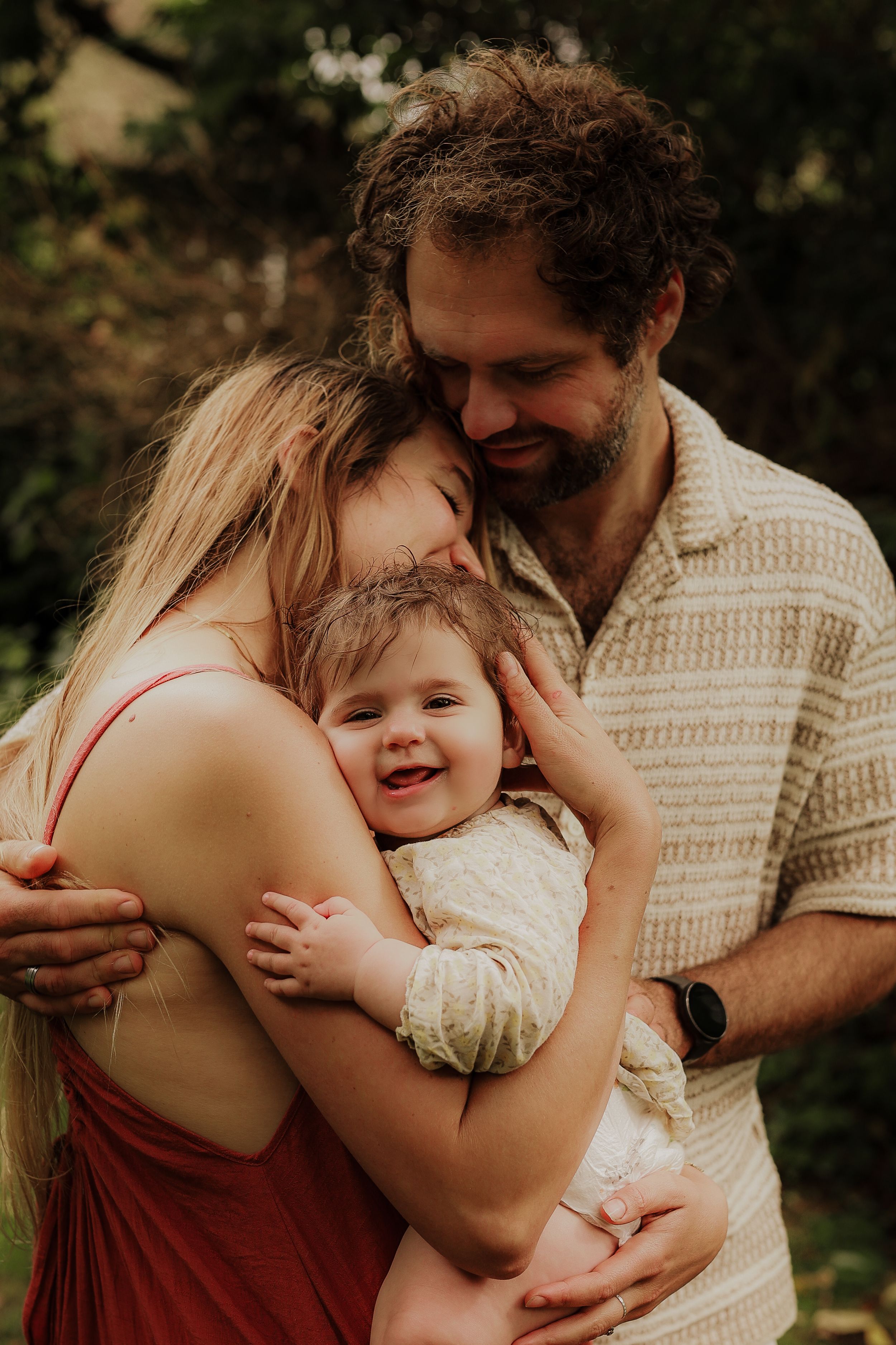 Loving parents embracing their smiling baby together in a warm outdoor family moment.
