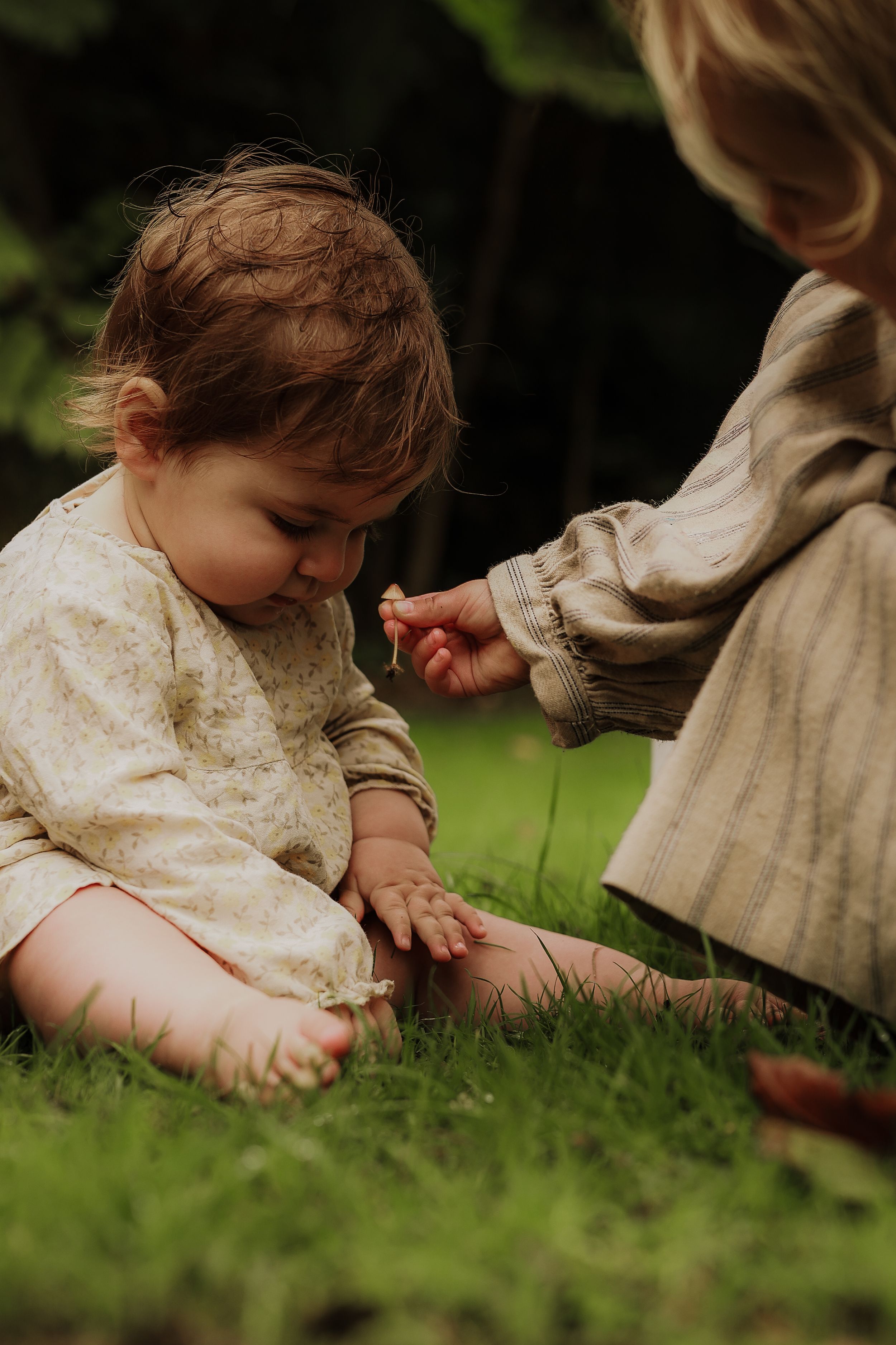 Curious baby sitting on grass while an older sibling offers a small flower outdoors.