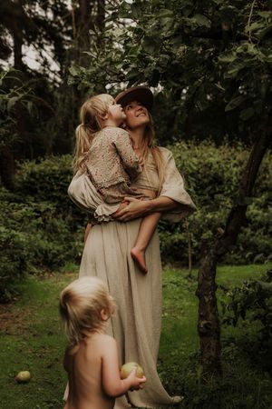 Mother in boho dress holding child near apple tree while toddler plays.