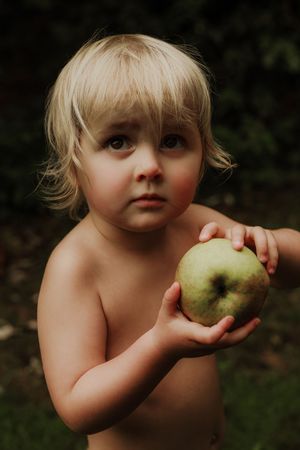 Blonde toddler holding a green apple outdoors in a garden setting.