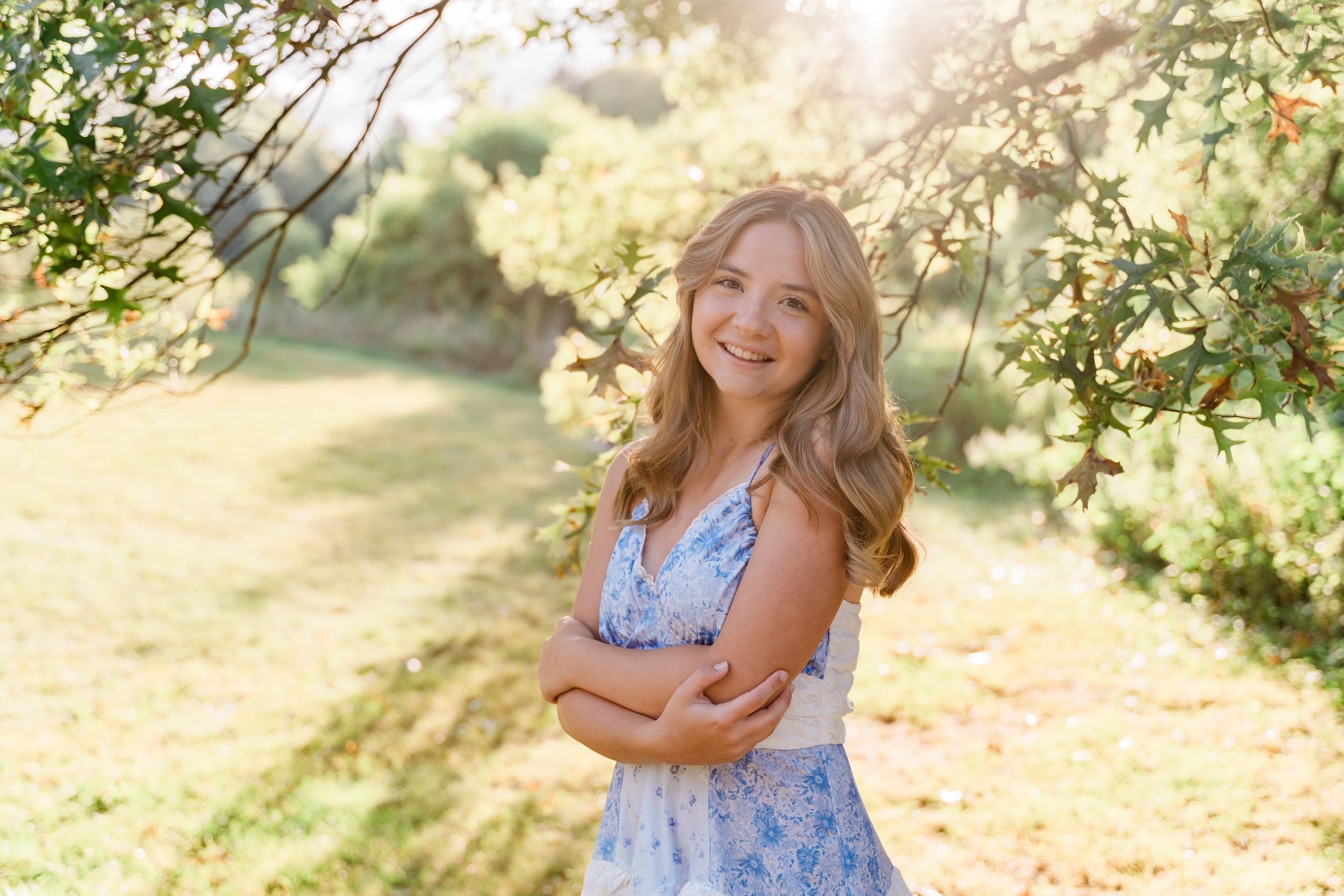 A person in a blue floral dress poses outdoors in a sunlit garden setting with blooming trees and natural lighting.