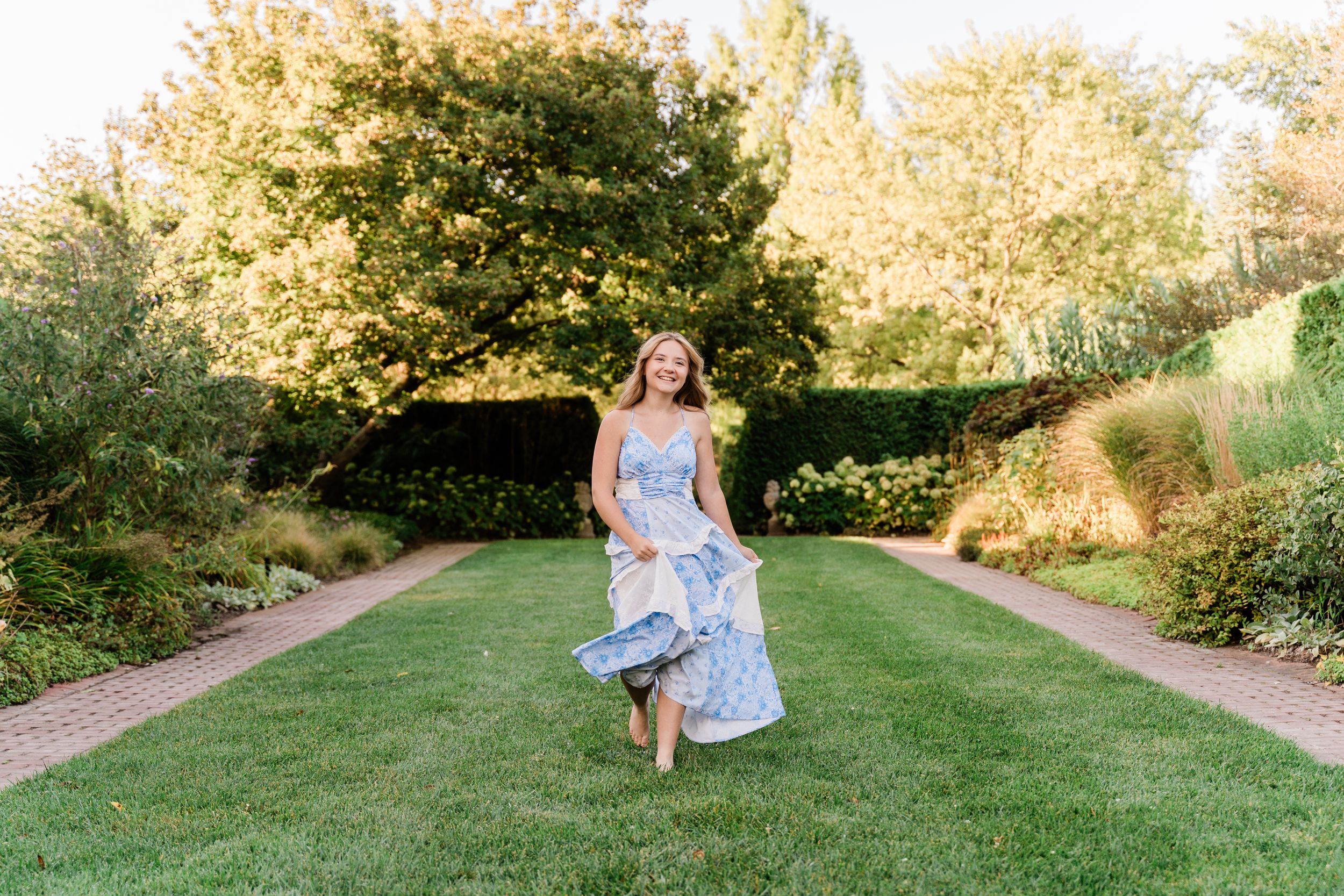 A person in a flowing blue floral dress twirls on a manicured lawn path surrounded by lush garden landscaping.