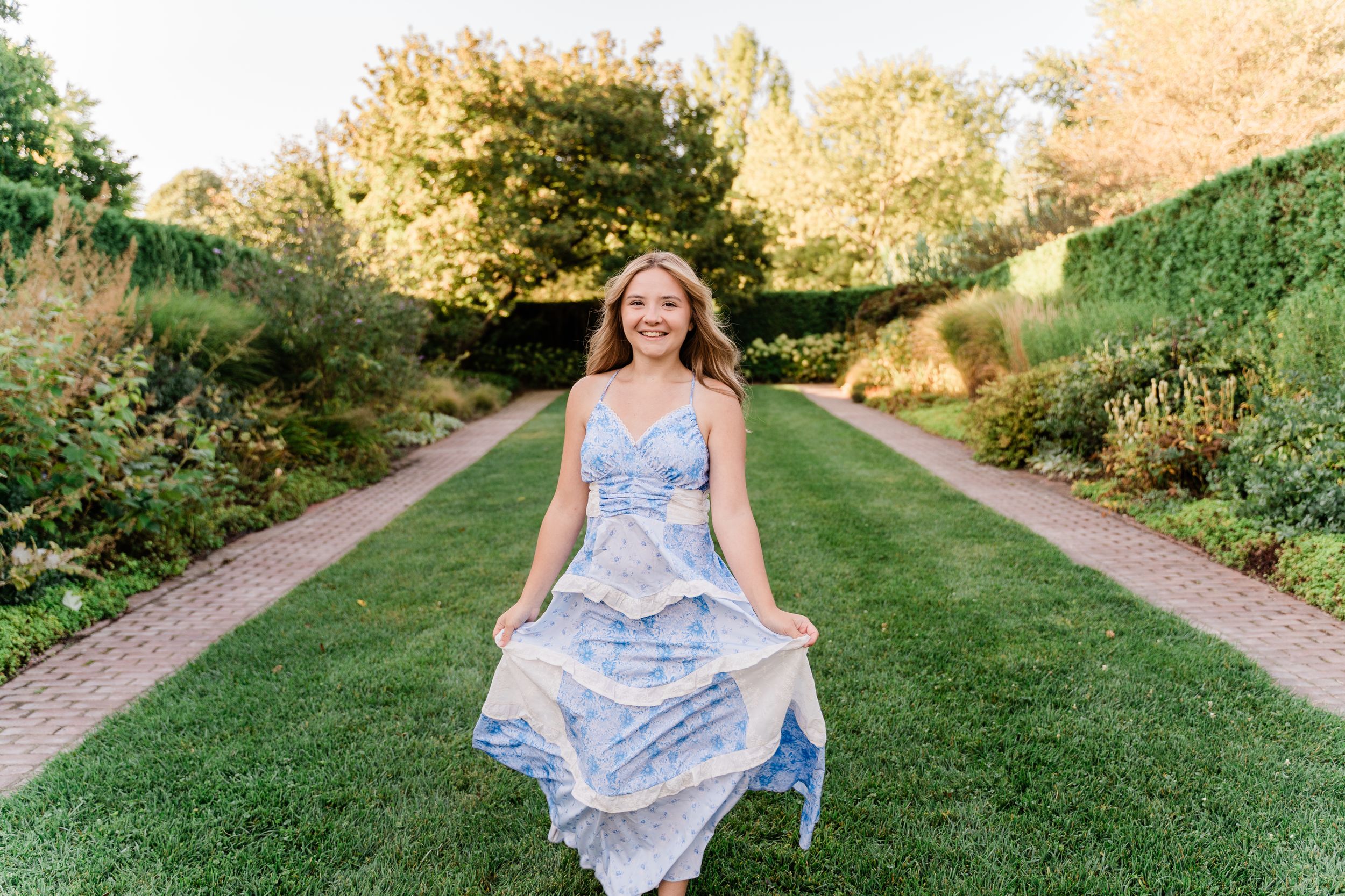 A figure in a flowy blue and white floral dress twirls on a sunlit garden path during golden hour.