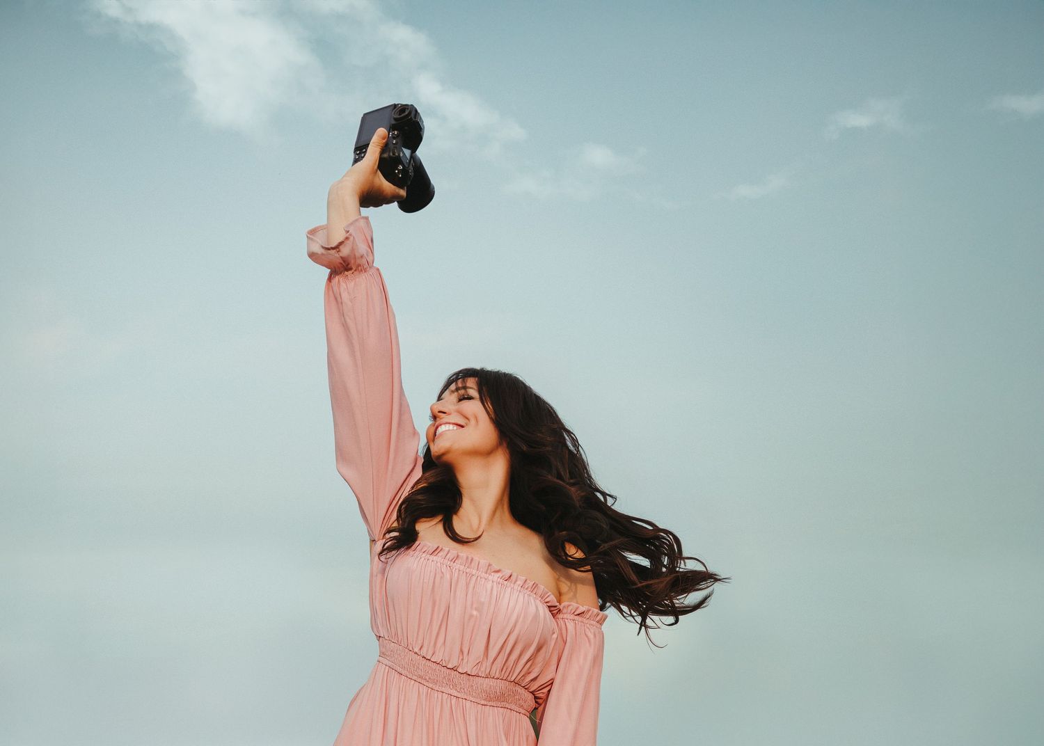 Woman in pink dress twirling joyfully against a cloudy blue sky background.