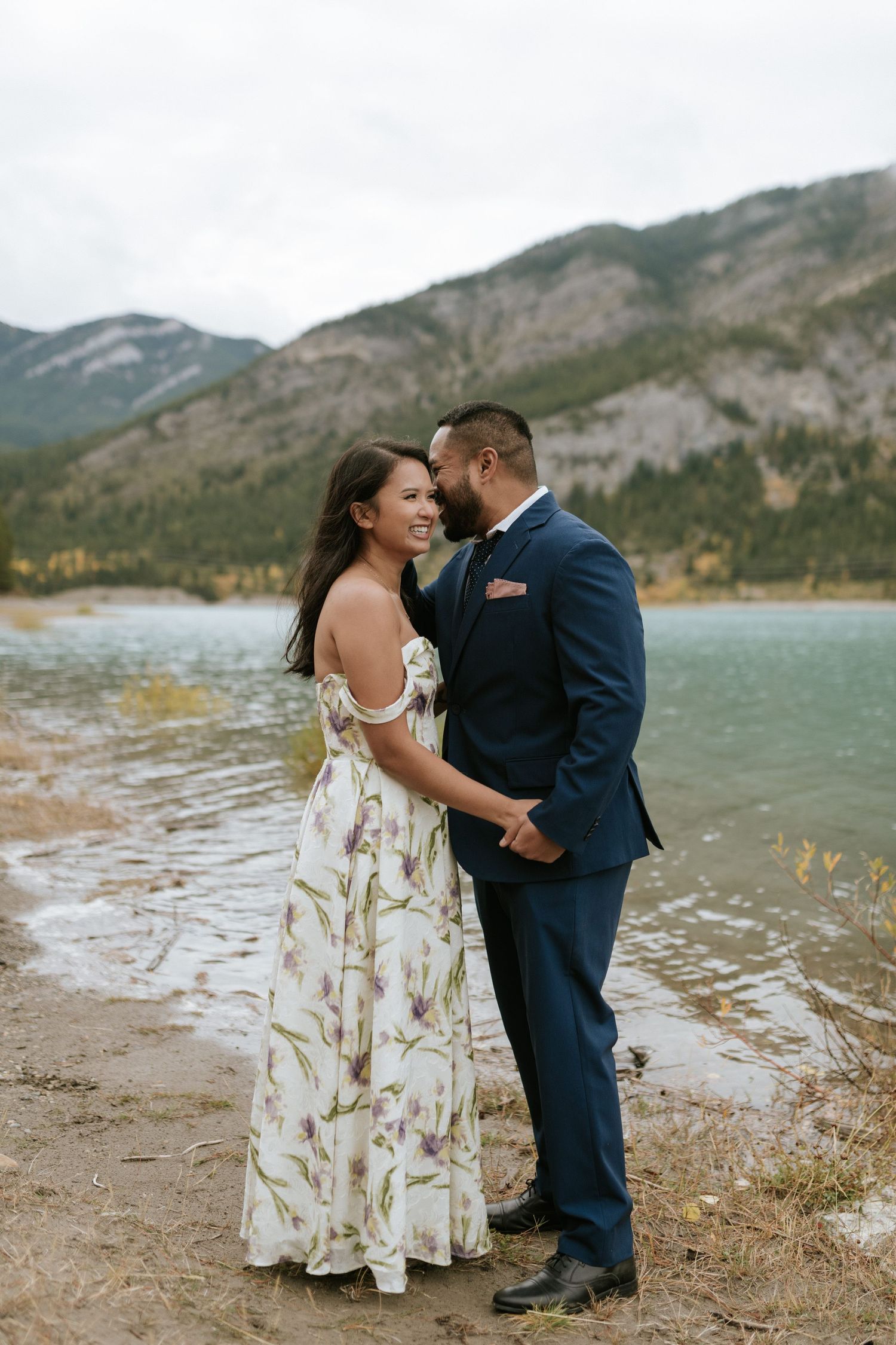 A couple embraces on a scenic mountain shoreline with turquoise waters and rugged peaks in the background.