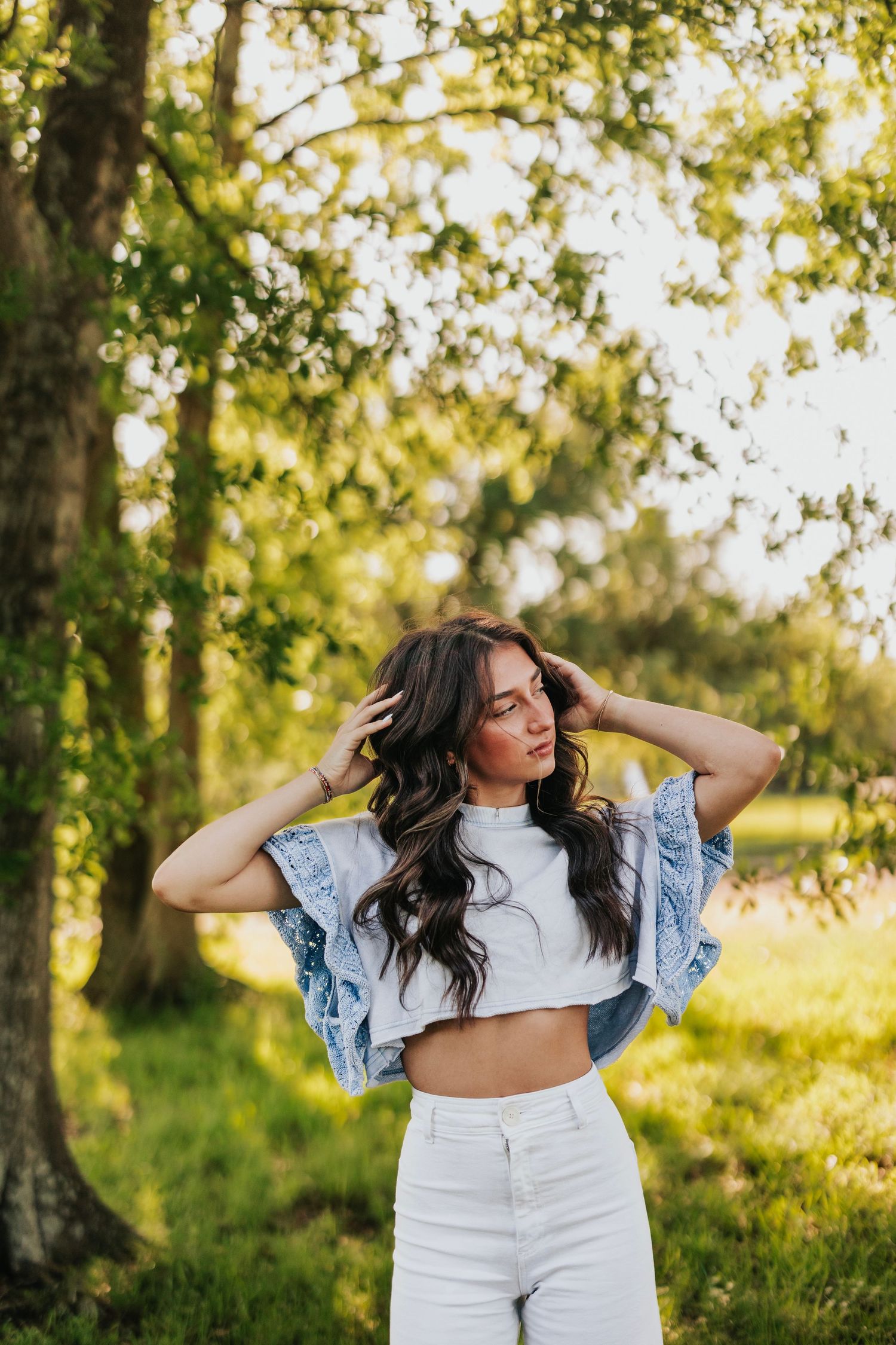 Woman in crop top and denim jacket poses in sunlit forest setting.