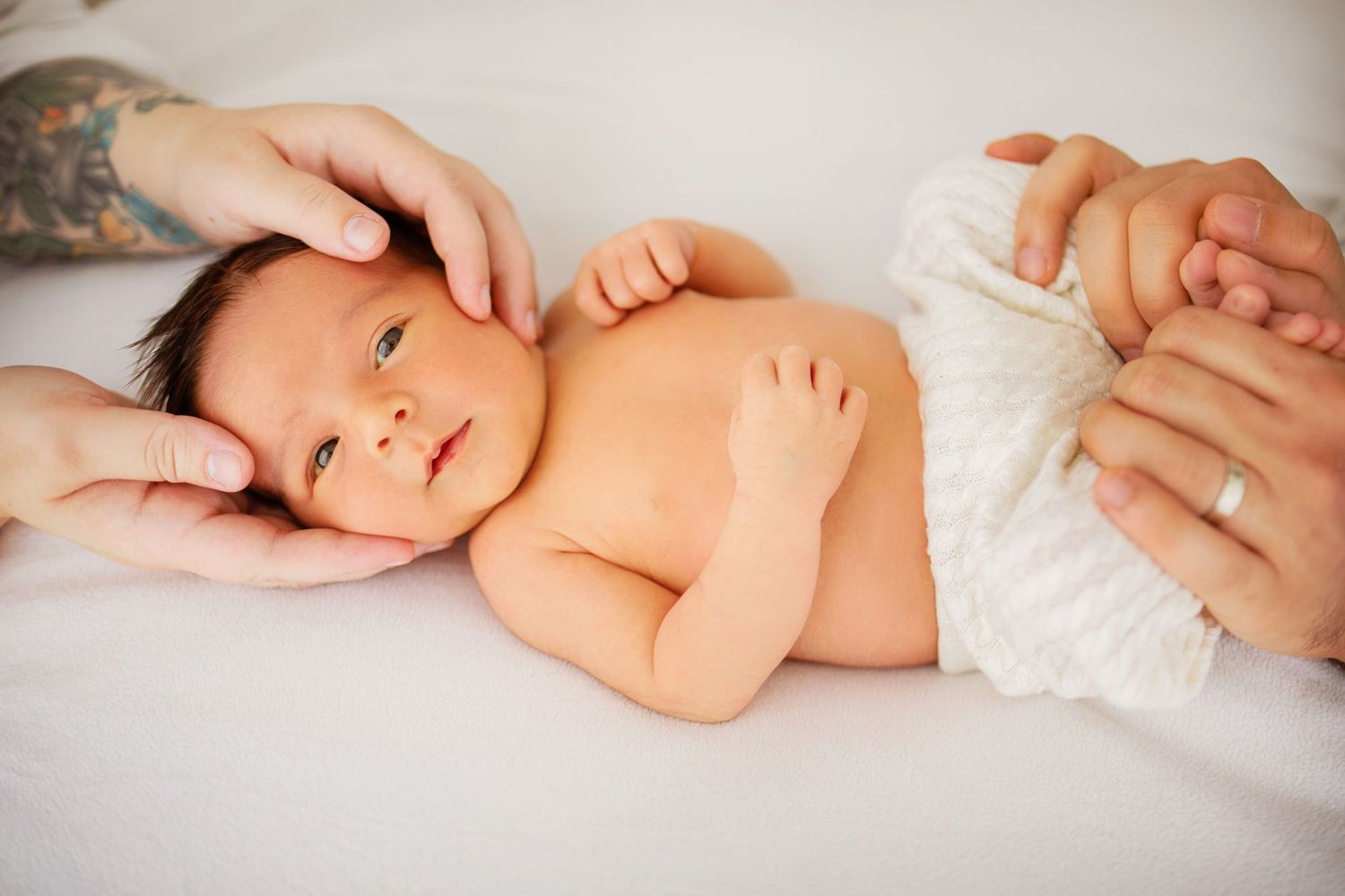 En nyfödd baby ligger lugnt på vita sängkläder medan milda händer ömt tar hand om dem.