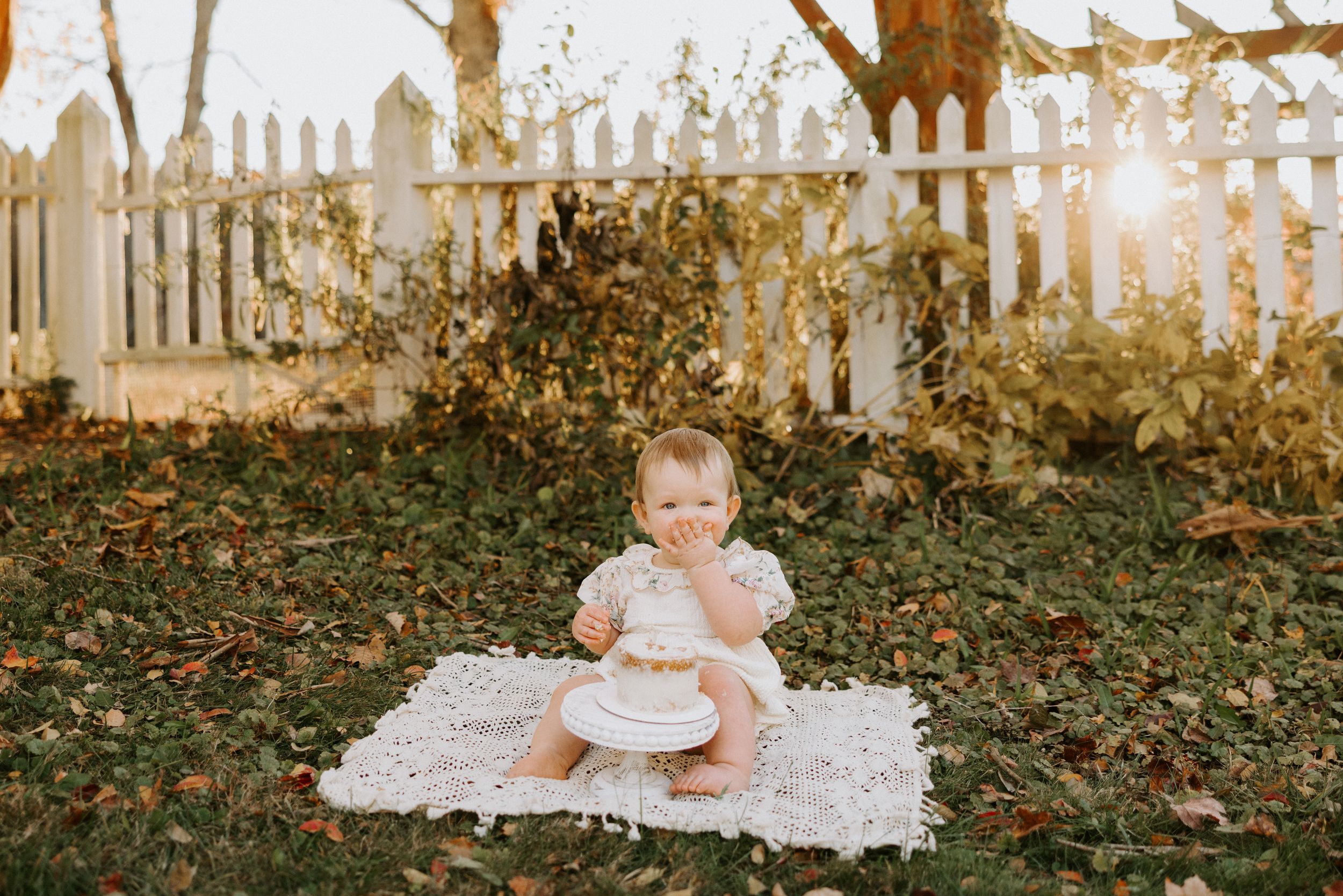 A baby sits on a white blanket in an autumn garden setting with a rustic white fence in the background.