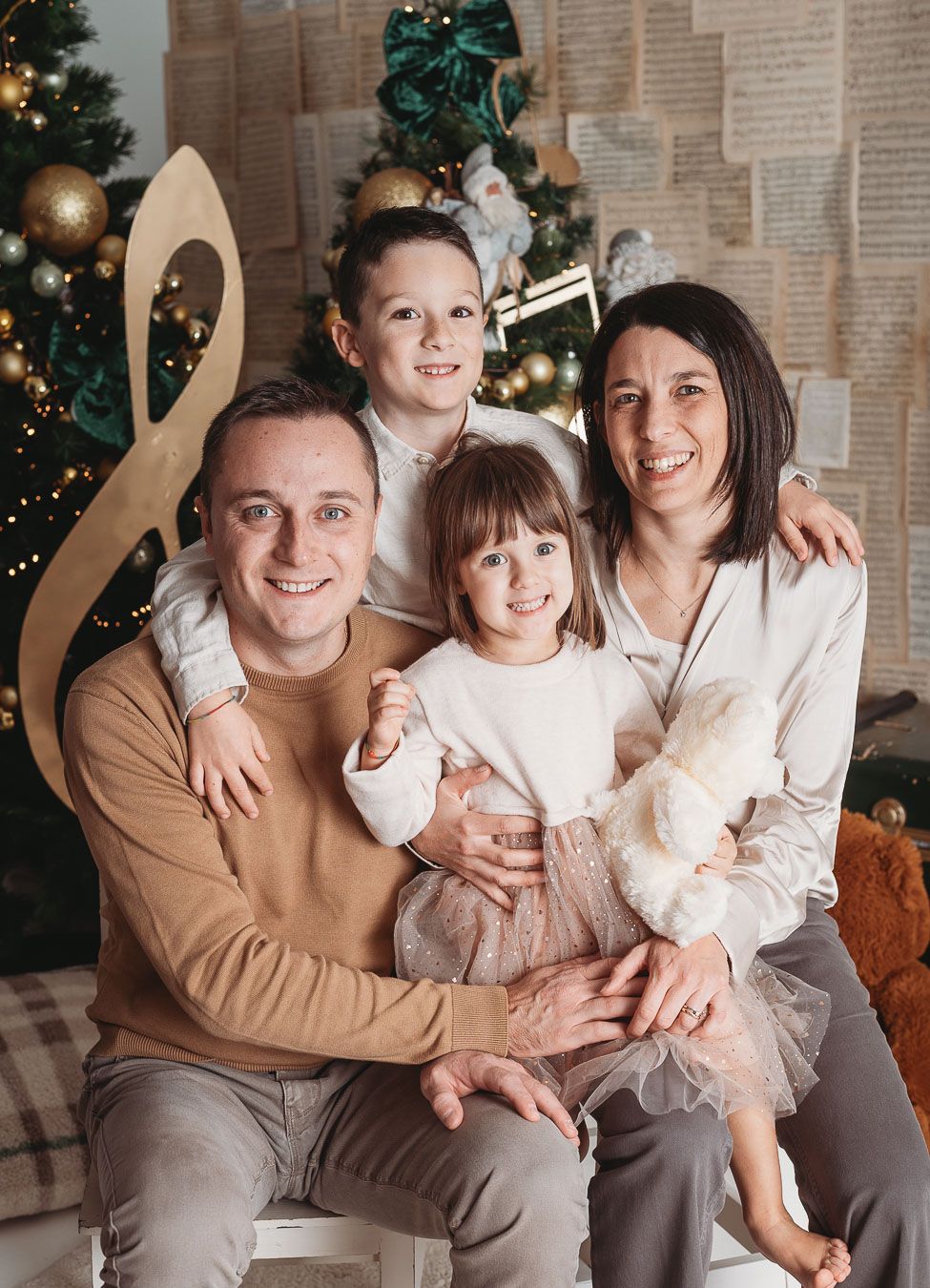 A family poses together on a couch in front of a decorated Christmas tree in matching cream and brown outfits.