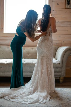 Two women in formal dresses, one in teal and one in white lace, stand together in a rustic wooden room.