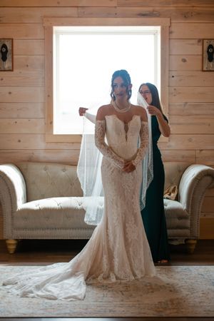 A bride in a white lace wedding dress stands in a sunlit room with wooden walls and vintage furniture.