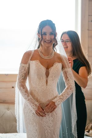 A bride in a detailed lace wedding gown shares a joyful moment with attendants in a sunlit room.