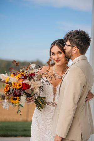Wedding couple embracing in a field with vibrant sunflower and autumn-colored bouquet.