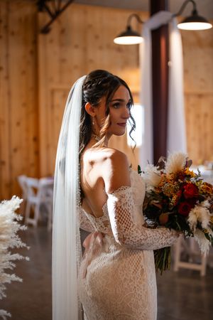 Bride in lace gown holds vibrant fall bouquet while posing in rustic wooden venue.