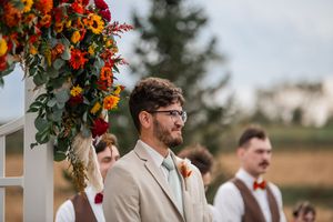 Wedding party stands under floral arch with autumn-colored decorations against outdoor landscape.