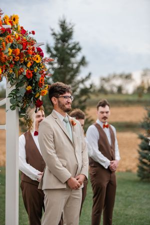 Groomsmen in tan and brown suits stand at outdoor ceremony with bright orange and yellow floral arrangements.