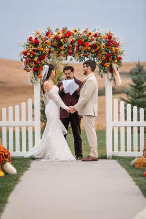 Outdoor wedding ceremony setup with white picket fence arch adorned with colorful fall flowers.