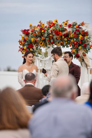 An outdoor wedding ceremony under a floral arch decorated with vibrant red and orange blooms.