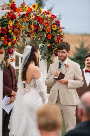 Wedding ceremony under floral arch with bright sunflowers and red blooms against white picket fence.