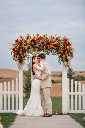 A romantic wedding moment under an arch adorned with bright autumn flowers and white picket fence details.