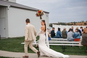 A wedding couple walks hand in hand across a grassy field near a white building.