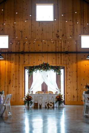 Rustic barn wedding venue interior with wooden walls, white drapes, and 'Mr & Mrs' sweetheart table decorated with greenery.