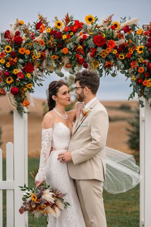 Wedding arch decorated with sunflowers and red roses against white picket fence backdrop.