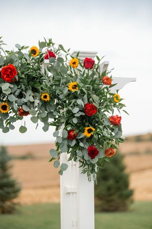 White wedding arch decorated with vibrant sunflowers, red roses, and eucalyptus greenery against a rural landscape.