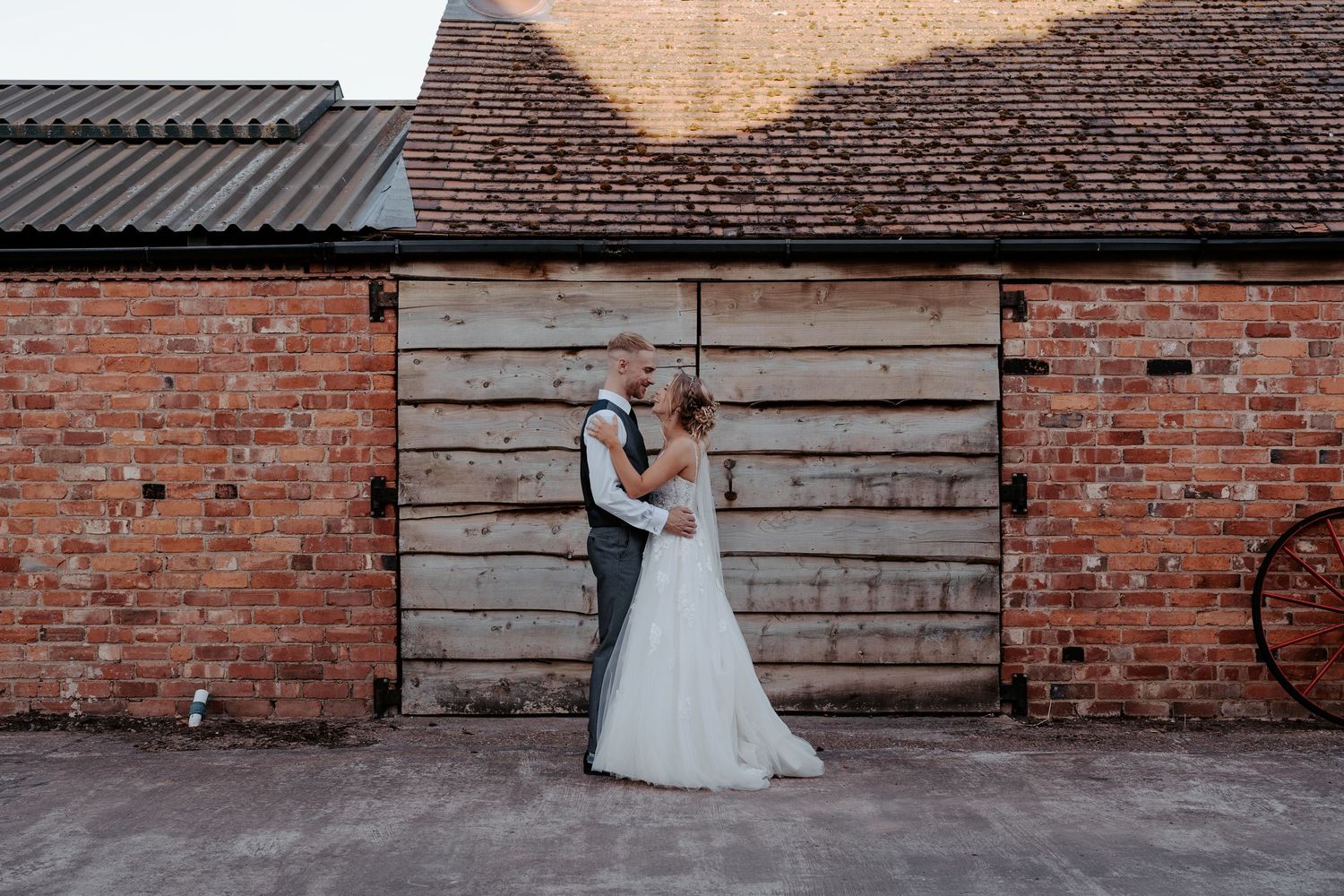 A couple stands in elegant wedding attire beneath a rustic brick archway with wooden doors at a historical venue.
