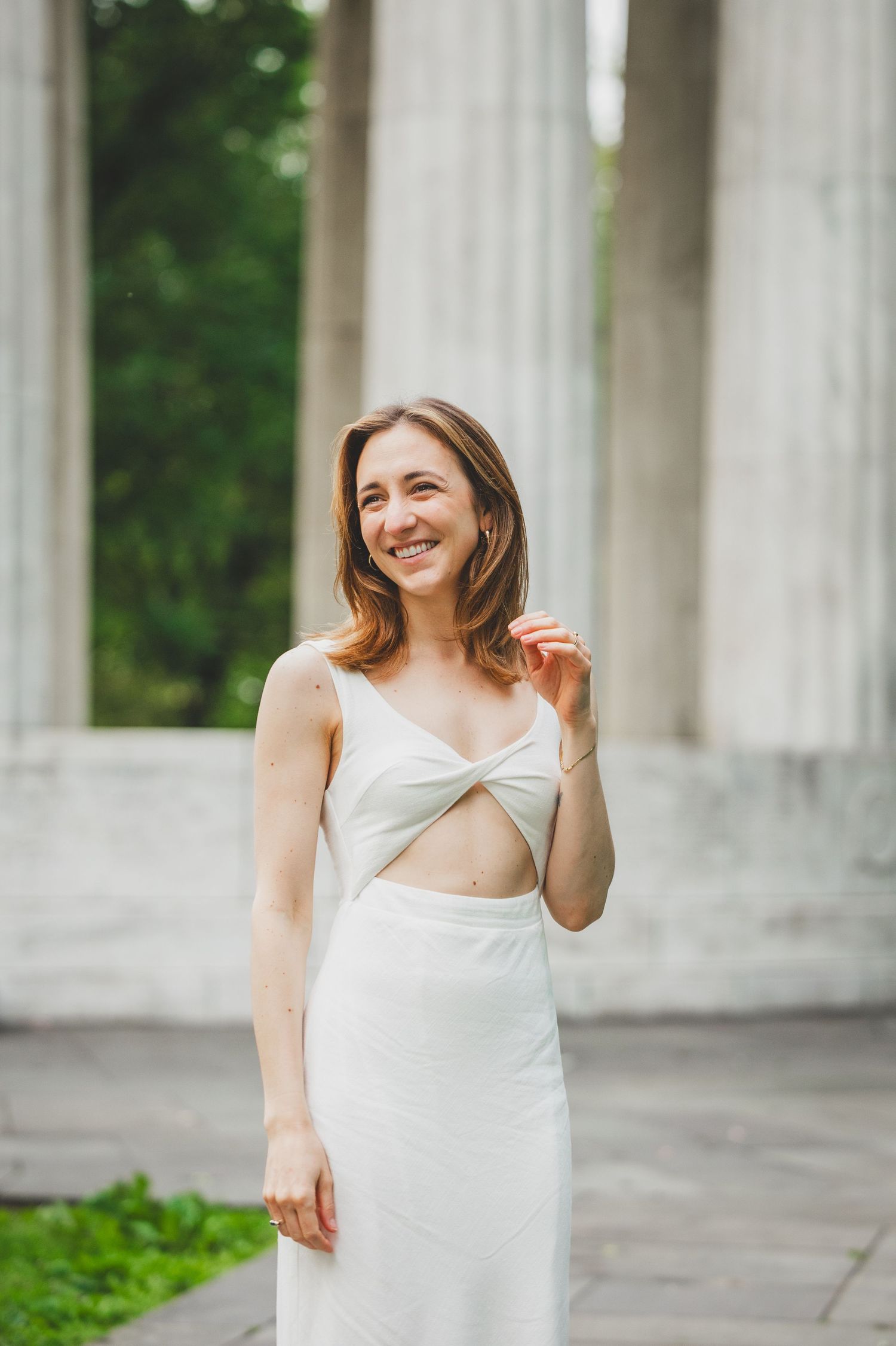 A person in a white dress poses gracefully between classical stone columns in an outdoor setting.