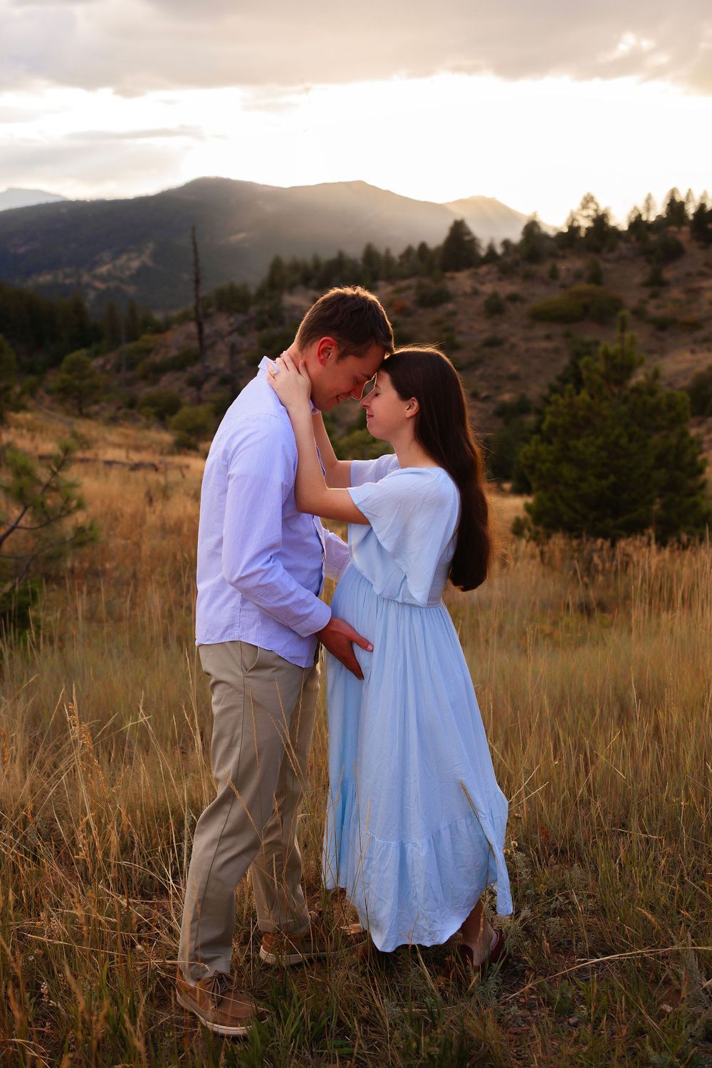 A couple shares a romantic moment in a field at sunset with mountains in the background.  Woman is pregnant.  Man has his hands on her bump