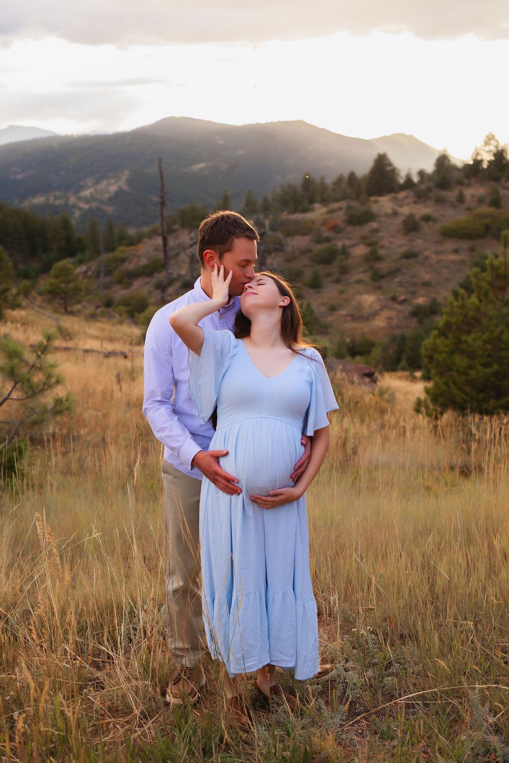 Couple in matching blue outfits share tender moments during sunset maternity photoshoot in a mountain meadow.