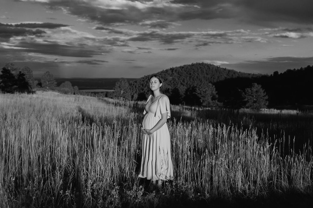 A dramatic black and white photo showing a pregnant woman in a flowing dress stood with her eyes closed in a tall grass field at sunset.