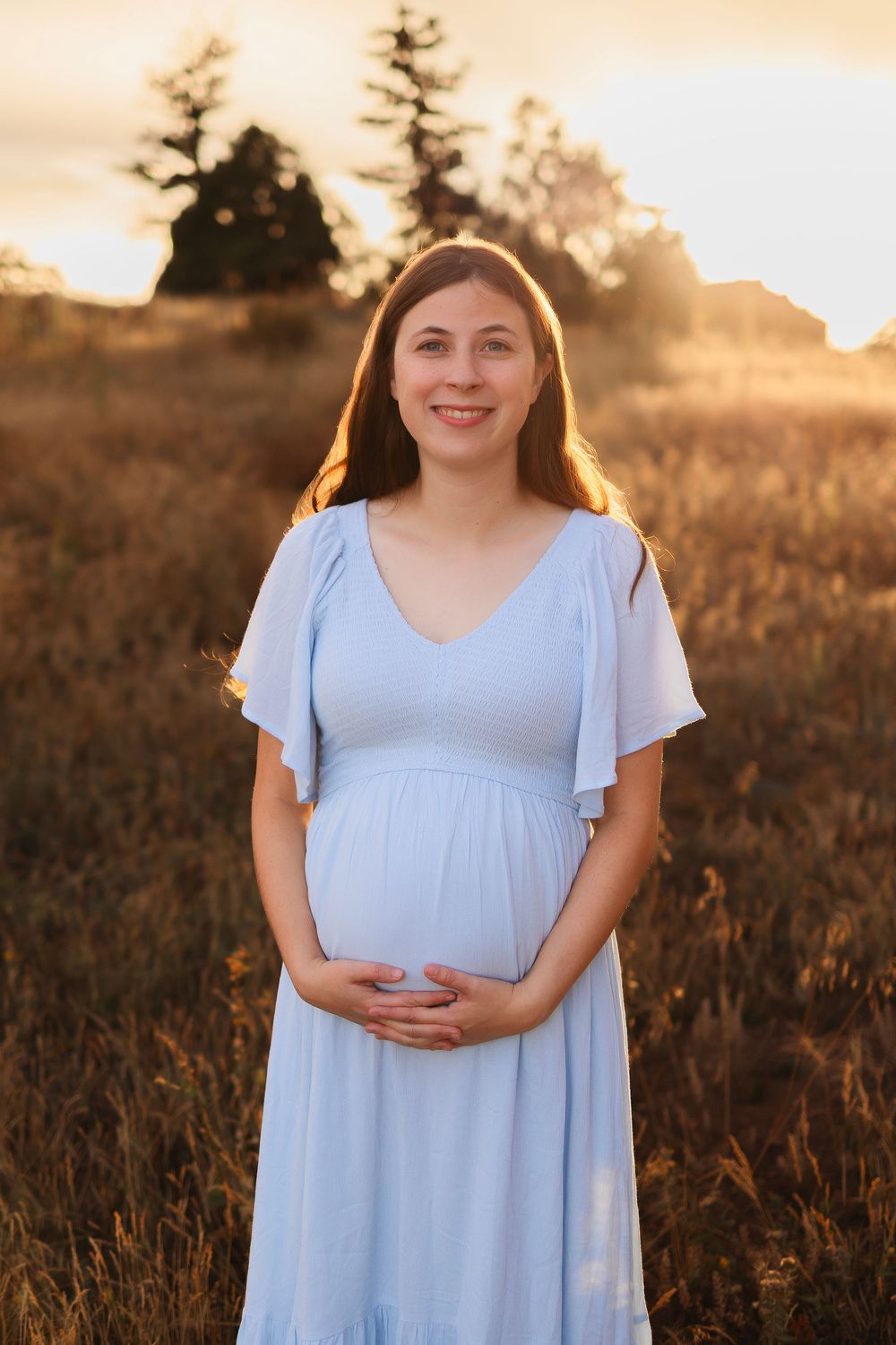 A pregnant woman in a flowing light blue dress stands in a golden field at sunset with trees silhouetted in the background.