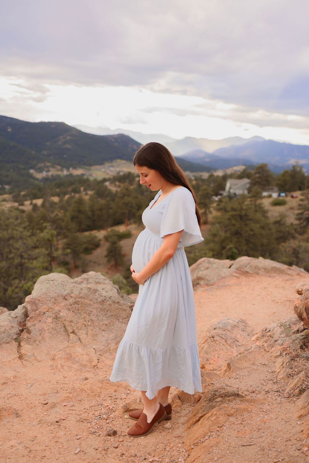 A pregnant woman in a flowing white dress stands on a rocky mountain overlook with scenic mountain ranges in the background.