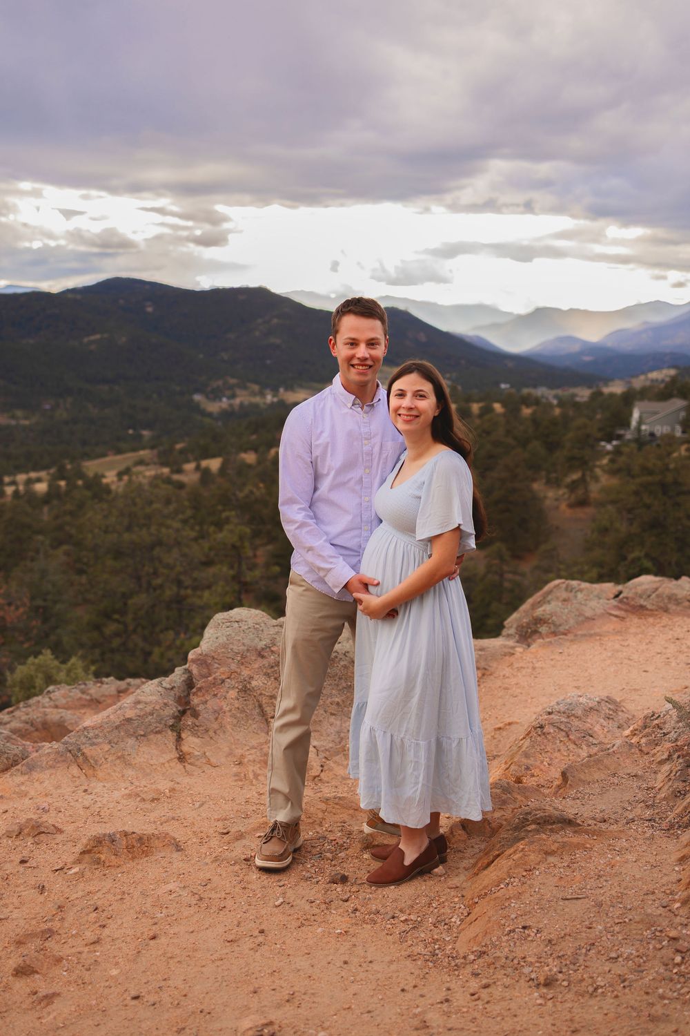 A couple stands together on a scenic red rock overlook with mountain views in the distance.  Maternity photoshoot mt falcon, colorado