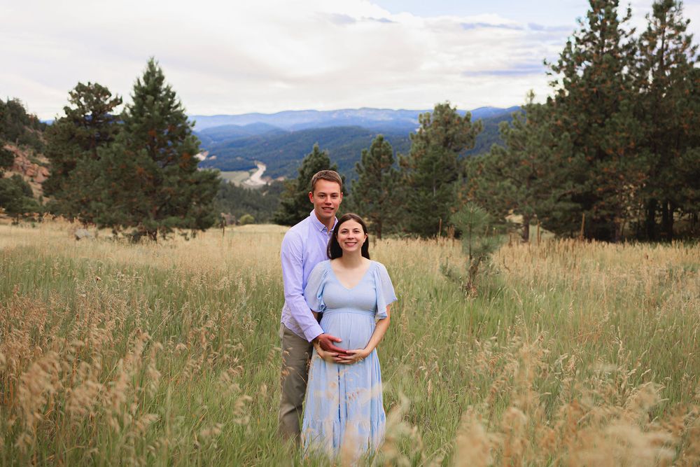 Couple shares romantic moments in a scenic mountain meadow with evergreen trees and golden grass at sunset. Maternity photoshoot
