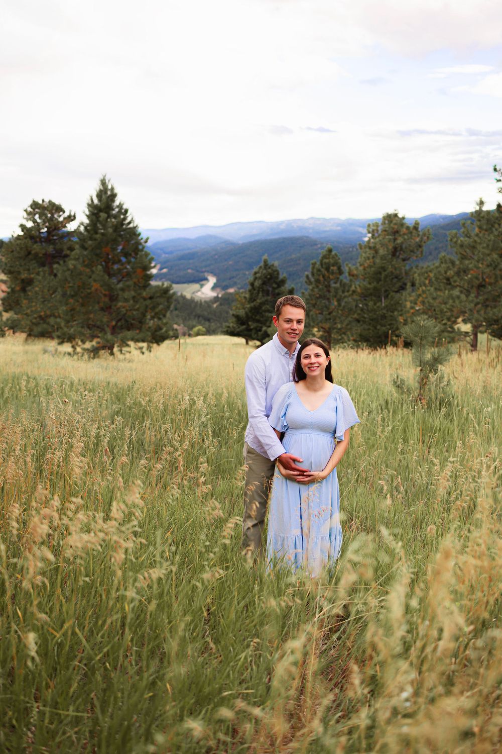 A couple stands together in a serene mountain meadow with tall grass and evergreen trees in the background.  Woman is pregnant.  Maternity
