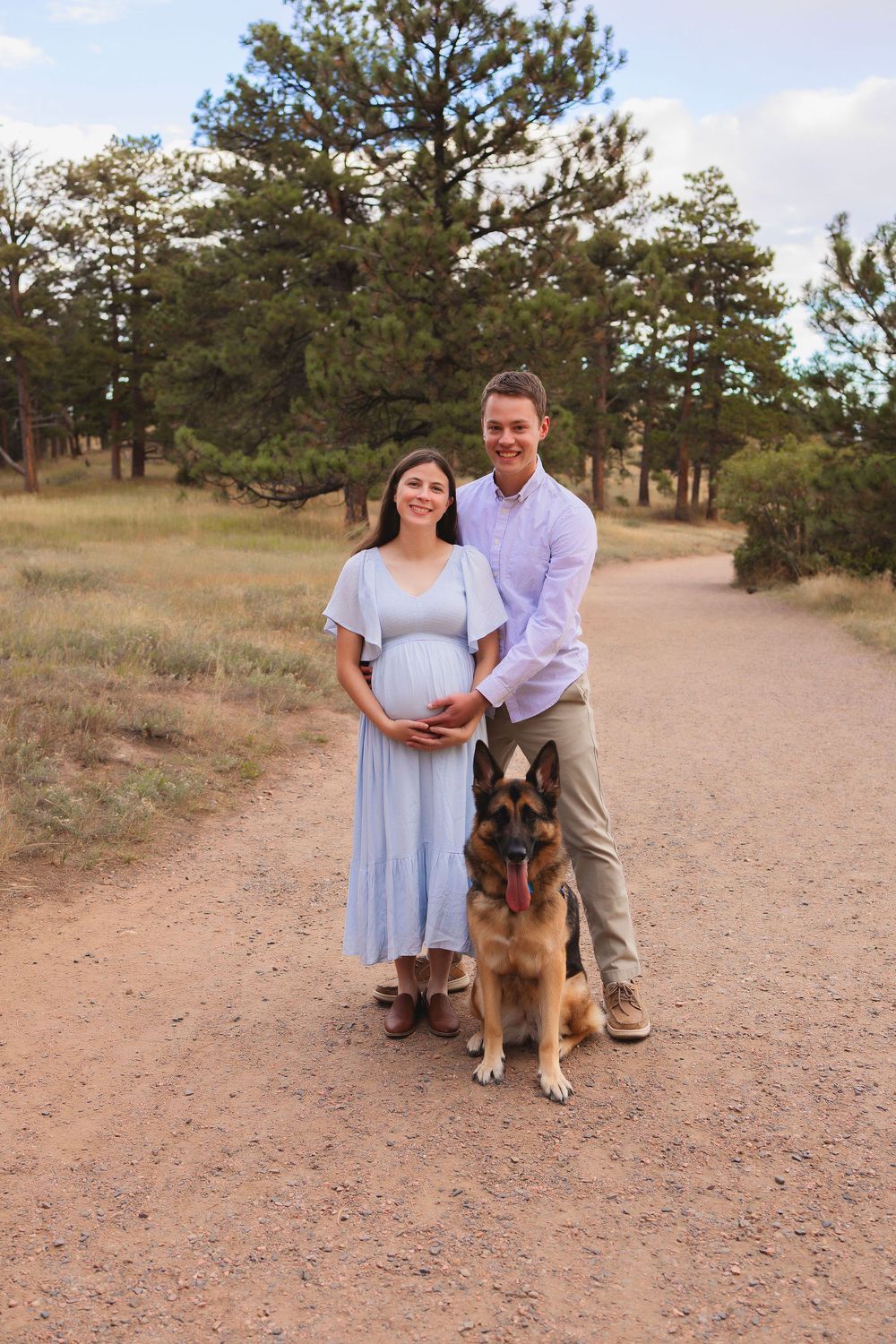 A couple and their German Shepherd dog pose together on a dirt trail surrounded by pine trees in a natural setting.  Woman is pregnant.