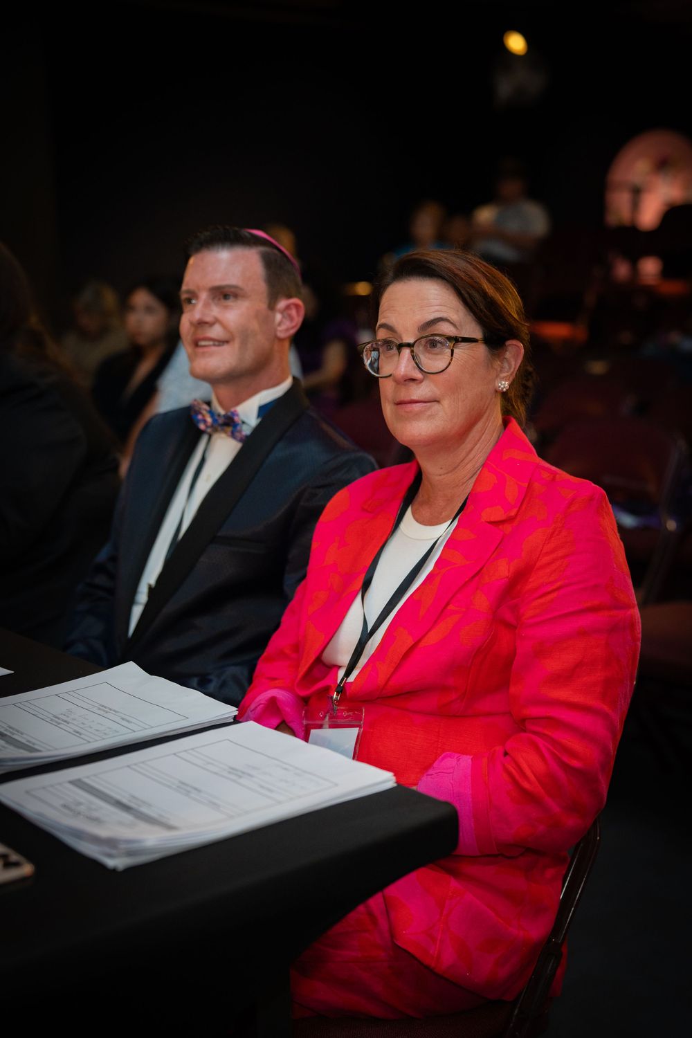 Person in bright red blazer sitting in audience at formal event with papers.