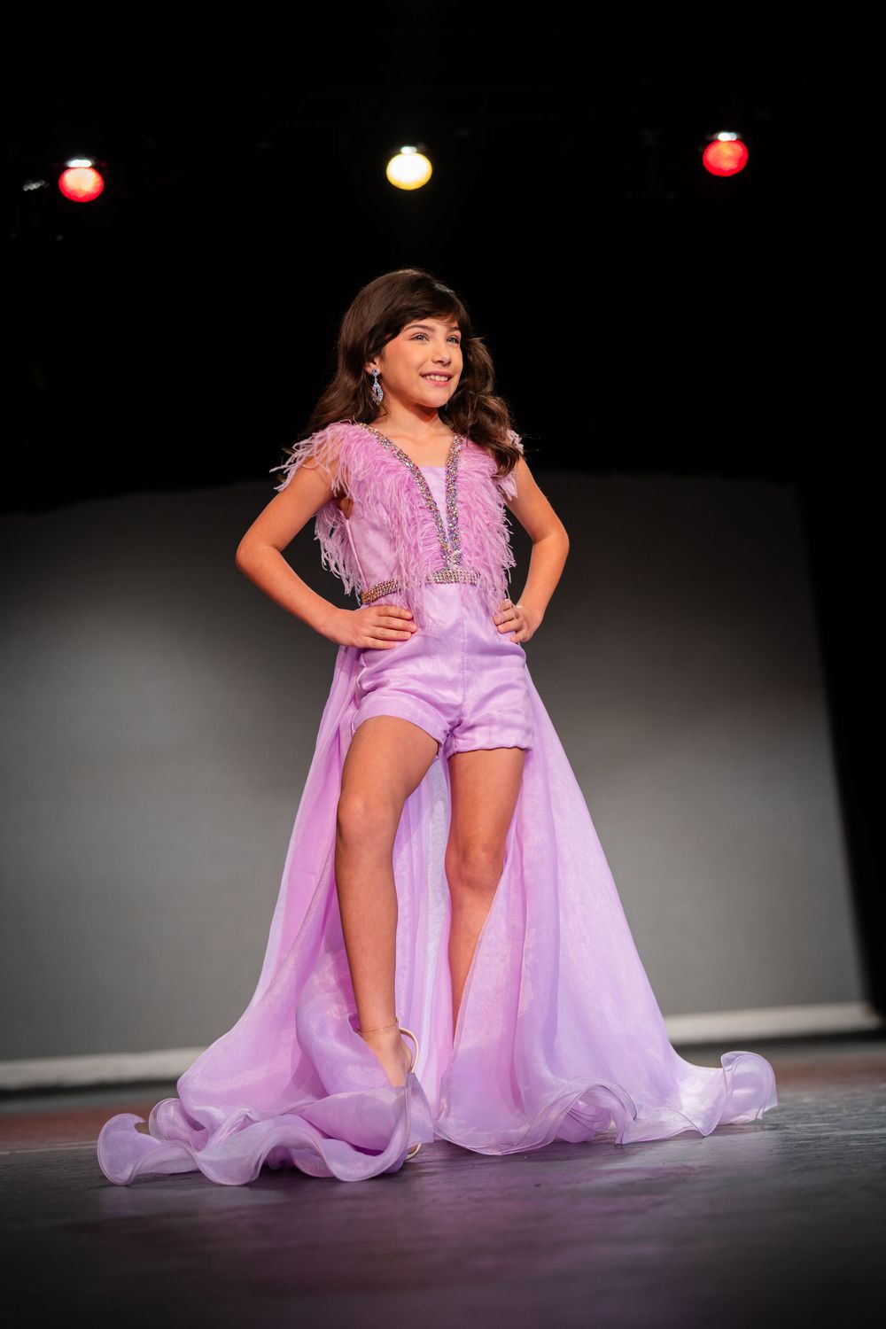 Performer in lavender costume doing expressive dance moves on dark stage.