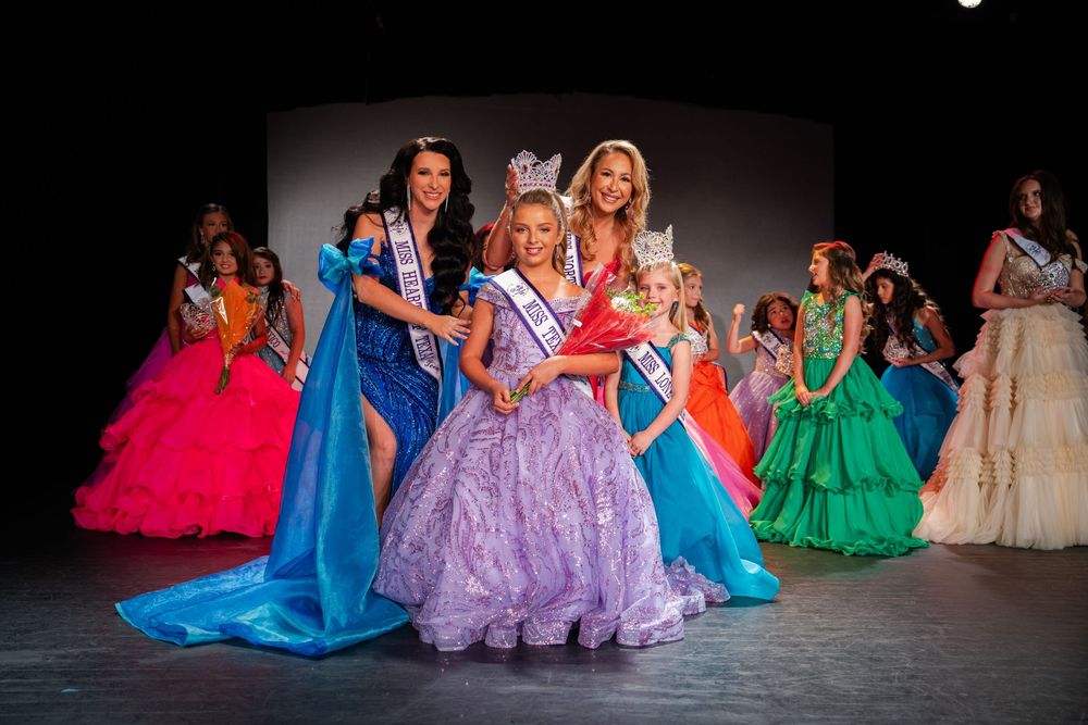 Wide shot of pageant contestants in colorful formal gowns lined up on stage.