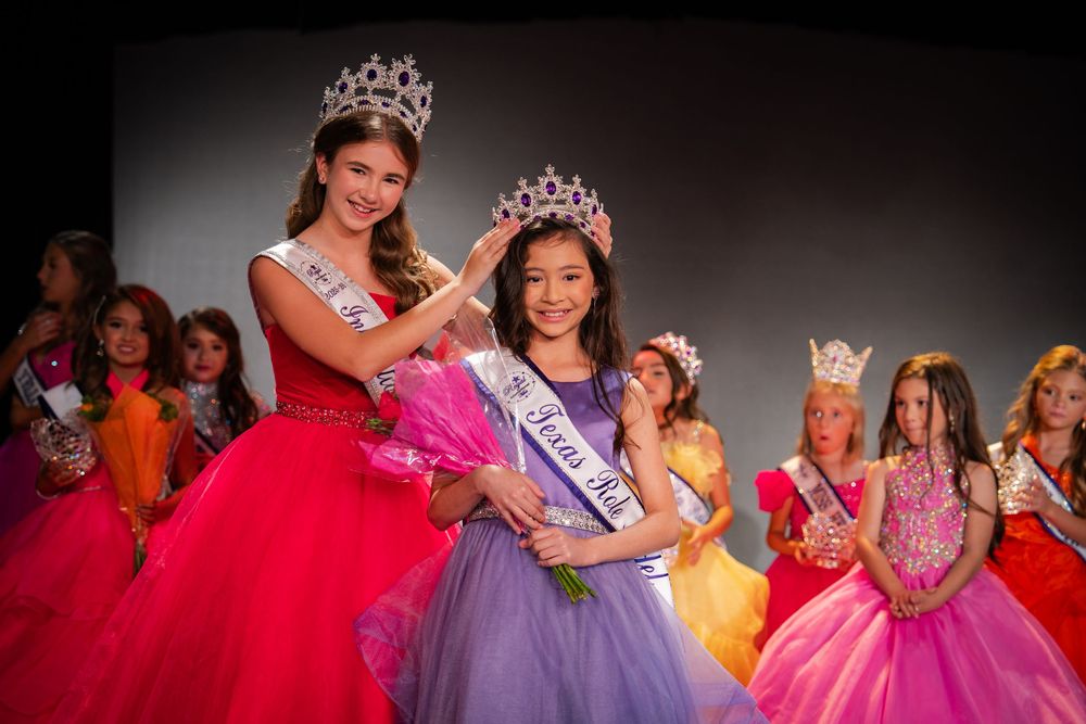 Row of pageant contestants wearing vibrant ball gowns and crowns line up on stage.